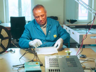 Ein freundlich lächelnder Mitarbeiter von VGS Lichttechnik sitzt an einem Arbeitstisch, bekleidet mit einem blauen Arbeitsoverall und weißen Handschuhen. In präziser Handarbeit setzt er die LED-Lichter in die ausgefrästen Module ein. Mit einem Lötkolben verbindet er sorgfältig die einzelnen LED-Stränge, wodurch die Grundlage für die Beleuchtung des Leuchtschildes geschaffen wird.